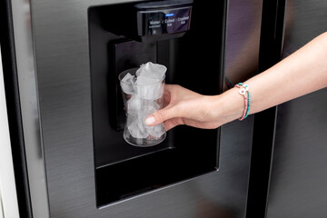 A woman stands in front of a sleek, stainless steel refrigerator, holding a glass filled to the brim with perfectly cubed ice - Powered by Adobe