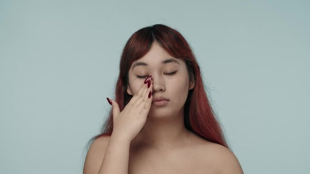 Close-up isolated slow motion video of a young seminude woman with red dyed hair and nude makeup looking straight at the camera, applying moistruizer, cream on her cheeck.