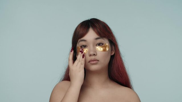 Close-up isolated slow motion video of a young seminude woman with red dyed hair and nude makeup fixing and patting eyepatches she applied under her eyes.