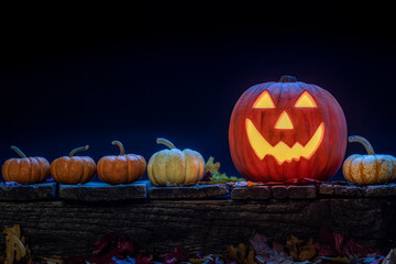 A pumpkin carved into a smiling Jack O Lantern sitting on an old wooden porch. There small pumpkins and fallen leaves as a Halloween decoration.