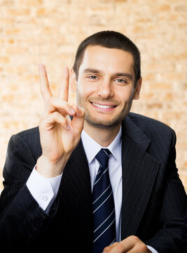 Happy smiling successful gesturing business man showing two fingers at office brown bricks wall background.
