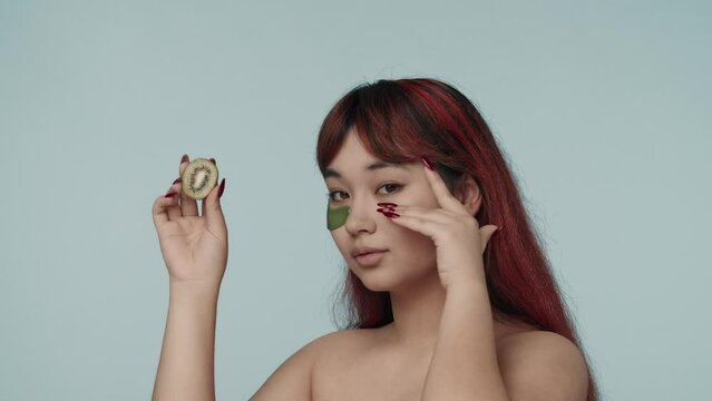 Close-up isolated slow motion video of a young seminude woman with red dyed hair and nude makeup wearing an eyepatch, holding a cut kiwi, touching her cheeck.