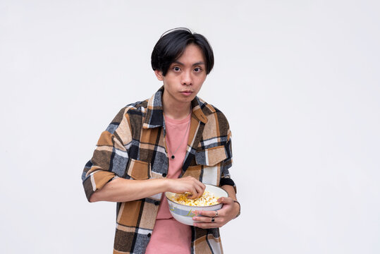 A young asian man looking spooked while holding a bowl of popcorn. Isolated on a white background.
