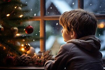 Fototapeta premium Cute little boy sitting by the window on Christmas eve. Child looking at the Christmas tree outside. Celebrating holiday at home. Traditional festive season.