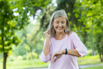 Senior gray-haired beautiful woman is running and doing sports in the park and checking the time on...