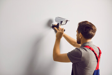 Portrait of a young technician man standing back installing surveillance CCTV camera on the wall indoors. Male worker using screwdriver while fixing security camera in modern office or at home. - Powered by Adobe