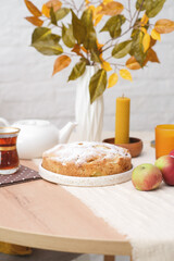 Autumn home decoration. Apple pie, Vase with autumn leaves branches, White teapot, candles on round wooden table on white background.