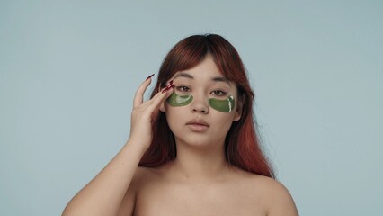 Close-up isolated photo of a young seminude woman with red dyed hair and nude makeup fixing and patting eyepatches she applied under her eyes. - obrazy, fototapety, plakaty