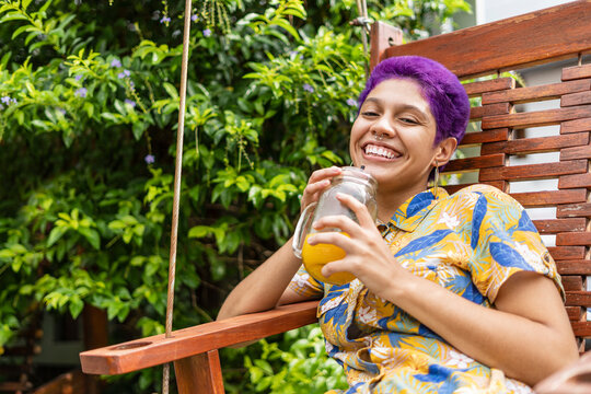non-binary gender woman sitting on a wooden chair outside a restaurant, very happy with a natural drink, smiling and looking at the camera with her short purple hair.