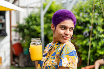 Portrait of a bisexual woman with short hair painted purple very happy and smilng with an orange juice in her hand outside a restaurant dressed in a yellow shirt.