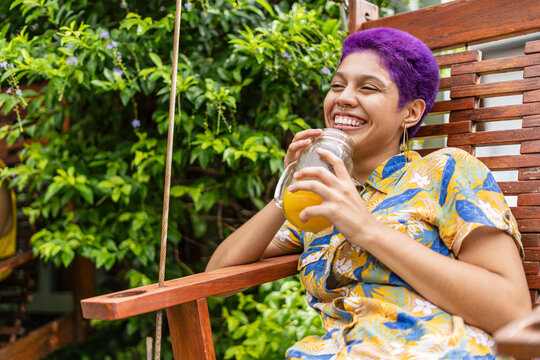 non-binary gender woman sitting on a wooden chair outside a restaurant, very happy with a natural drink, smiling and looking at the camera with her short purple hair.