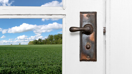 Close-up horizontal photo of an old vintage metal door lever on a white wooden door with windows. A green agricultural field, trees and blue skies with white clouds is visible through the window.