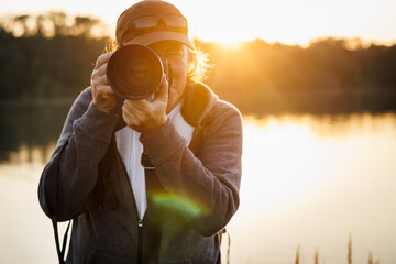 Wildlife photographer with camera photographing nature on lake at sunset. Front view of man, selective focus at lens - obrazy, fototapety, plakaty