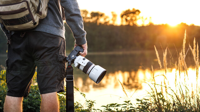 Landscape and wildlife photographer holding camera with telephoto lens and looking for composition at lake during sunset