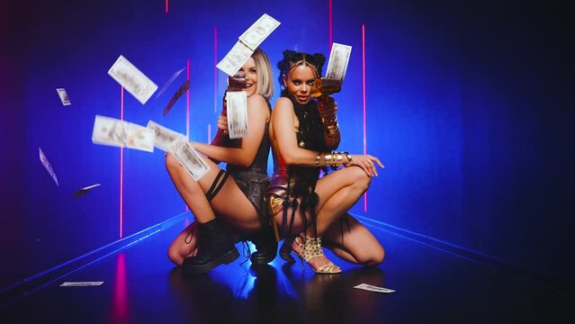 Two happy women shoot money from gun dollars fall fly hand holding pistol. Beauty rich sexy girl fashion model smiling face, woman blond hair short dress, studio neon red blue light 4k. joy fun party