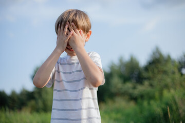 Children's emotions. Boy in light T-shirt stands on summer in nature, covered his face with hands.