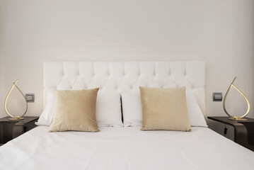Headboard in a modern bedroom with white capitone upholstered headboard, gold cushions and twin decorative lamps