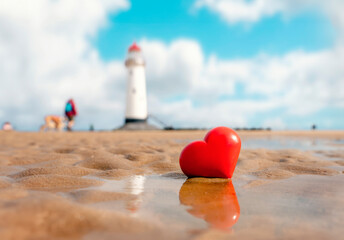 Fototapeta premium Big red heart in sand at coast against lighthouse. Fall in love with holiday at seaside concept