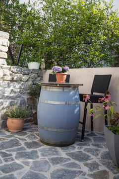 Vintage garden design:Old wooden wine barrel used as bar table with barstools on a terrace with virgin stone pavement in front of a virgin stone wall, surrounded by a tree, pots with flowers, lavender