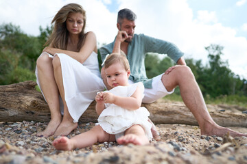 Fototapeta premium A young parents sitting in thoughts on the sand beach. A small baby playing in front.