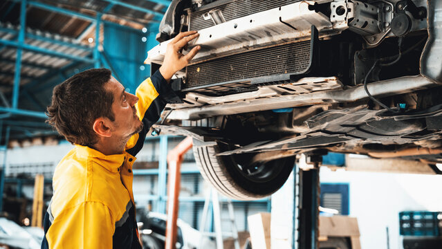 Vehicle mechanic conduct car inspection from beneath lifted vehicle. Automotive service technician in uniform carefully diagnosing and checking car's axles and undercarriage components. Oxus