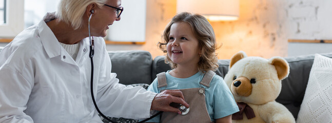 Concept of healthcare, medical assistance, insurance. Kind senior female paediatrician doctor examining little child, cute girl during home visit or clinic check up. Prescribing treatment, banner