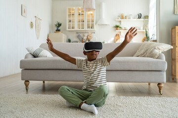 Overjoyed little African American boy child wearing vr headset enjoying spending leisure time in virtual reality. Happy kid sitting on floor in living room playing online game simulation, exploring AR