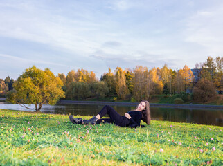 Naklejka na ściany i meble A beautiful girl poses while sitting on the grass by a pond in an autumn park.