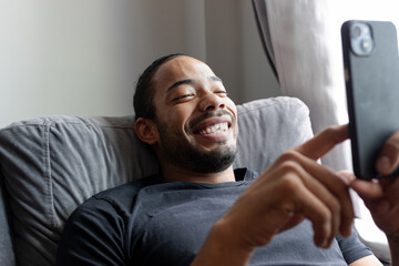 Dark-skinned man lying on the sofa at home while looking at the mobile phone