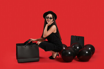 Beautiful young woman with shopping bags and balloons sitting on red background. Black Friday sale