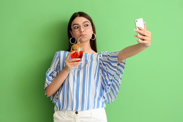 Beautiful young woman with glass of tasty aperol spritz taking selfie on green background