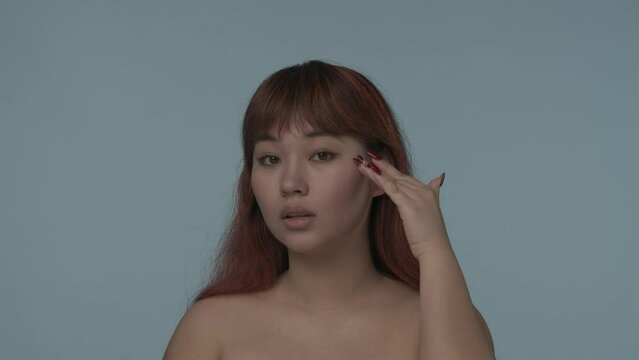 Close-up isolated slow motion video of a young seminude woman with red dyed hair and nude makeup looking at the camera, touching her cheeck and smiling. HDR BT2020 HLG Material.