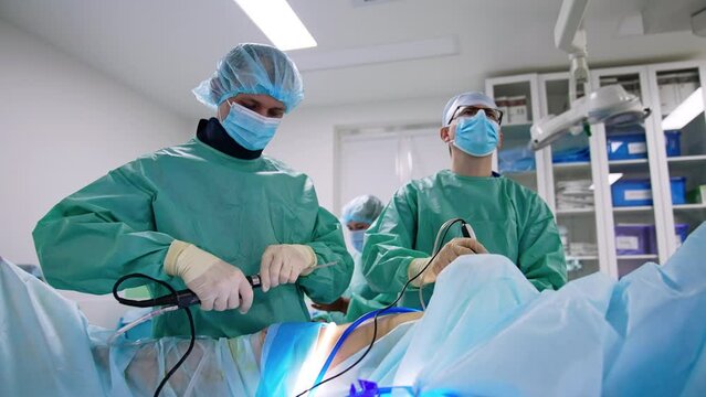 Male adult surgeon wearing glasses uses laparoscopic device. Assisting doctor prepares another tool. Low angle view.