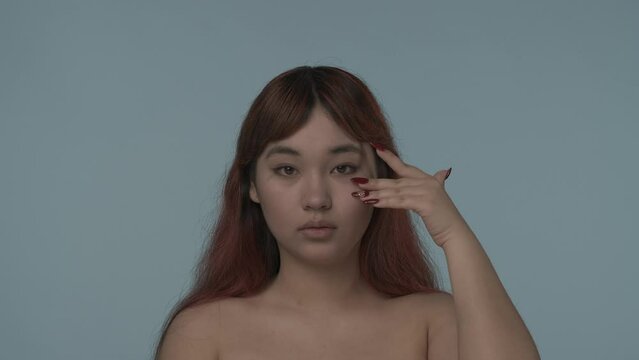 Close-up isolated slow motion video of a young seminude woman with red dyed hair and nude makeup looking at the camera, touching her cheeck and smiling. HDR BT2020 HLG Material.