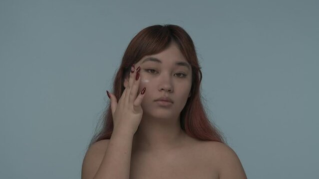Close-up isolated slow motion video of a young seminude woman with red dyed hair and nude makeup looking straight at the camera, applying moistruizer, cream on her cheeck. HDR BT2020 HLG Material.