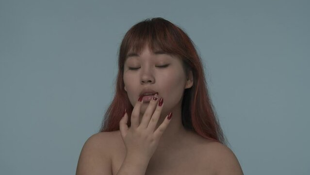 Close-up isolated slow motion video of a young seminude woman with red dyed hair and nude makeup applying a lip oil, lip balm on her lips. HDR BT2020 HLG Material.