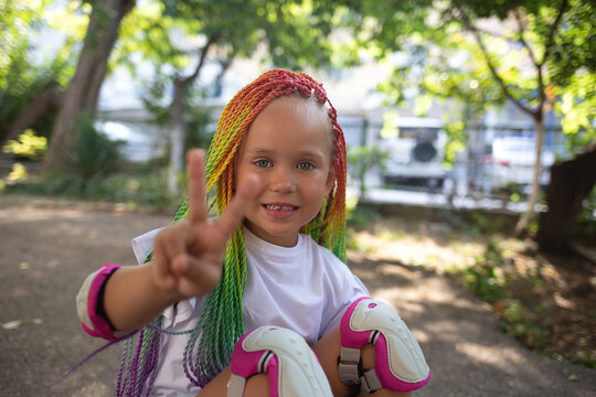 little cute girl with colorful afro pigtails sits on a skateboard and shows a peace sign