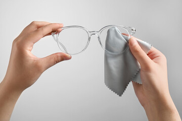 Woman wiping glasses with microfiber cloth on light grey background, closeup