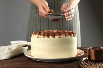 Woman dusting delicious tiramisu cake with cocoa powder at wooden table, closeup