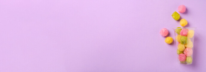 Obraz premium Plastic cup with color cotton balls on violet background, top view. Banner design with space for text