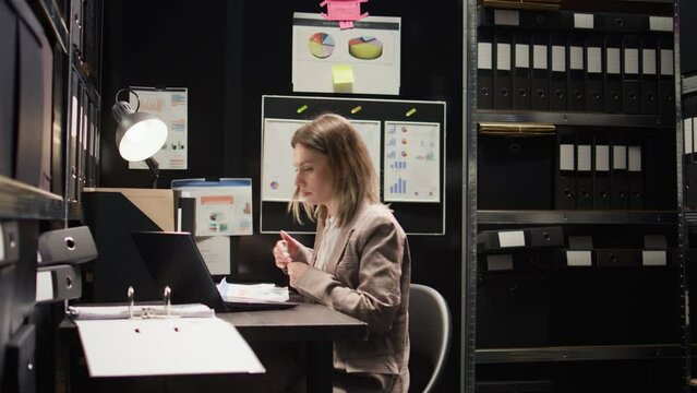 Focused female investigator conducting investigation and reviewing evidence. Zoom-in shot of inspector sitting at desk with laptop and case files for comparing statements and profile of the suspect.