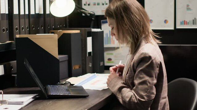 Policewoman captured in midst of investigation, examining records and evidence. Detailed view of officer at desk with laptop and case files opened for cross-referencing suspect profile and statements.