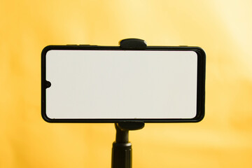 Landscape phone with white screen fixed to tripod on yellow background, for mockup design. - obrazy, fototapety, plakaty