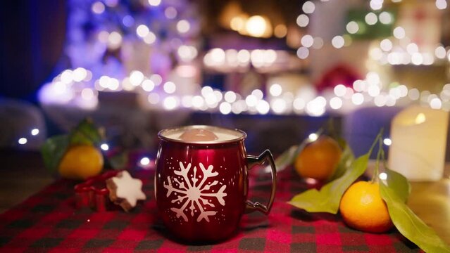 Close up dolly shot of red glossy cup with white snow flake pattern with hot cocoa and chocolate bomb floating on top haunted on table with red plaid napkin near fireplace. Winter leisure concept 4K