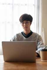Fototapeta premium A smart young Asian male college student is working on his project on a laptop in a cafe.