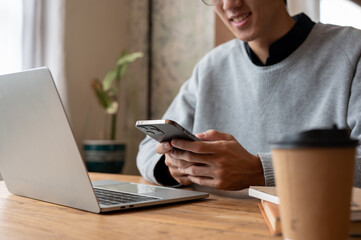 Close-up image of a handsome Asian man using his smartphone while working remotely at a coffee shop.