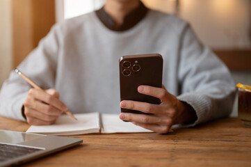 A male college student using his smartphone while doing homework at a table in a coffee shop.