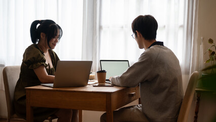 Two happy young Asian college students are working remotely at a coffee shop together, using laptop.