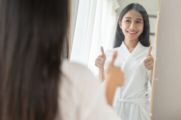 A confident woman in a bathrobe is smiling and giving herself a thumbs up in front of the mirror.