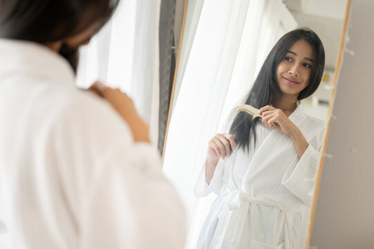 Beautiful lady in a bathrobe is combing her hair with a brush while standing in front of the mirror